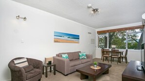 Ein Wohnzimmer mit einer Couch, einem Sessel, einem Couchtisch und einem Essbereich mit Stühlen. An der Wand hängt ein Strandbild und es gibt einen Deckenventilator.