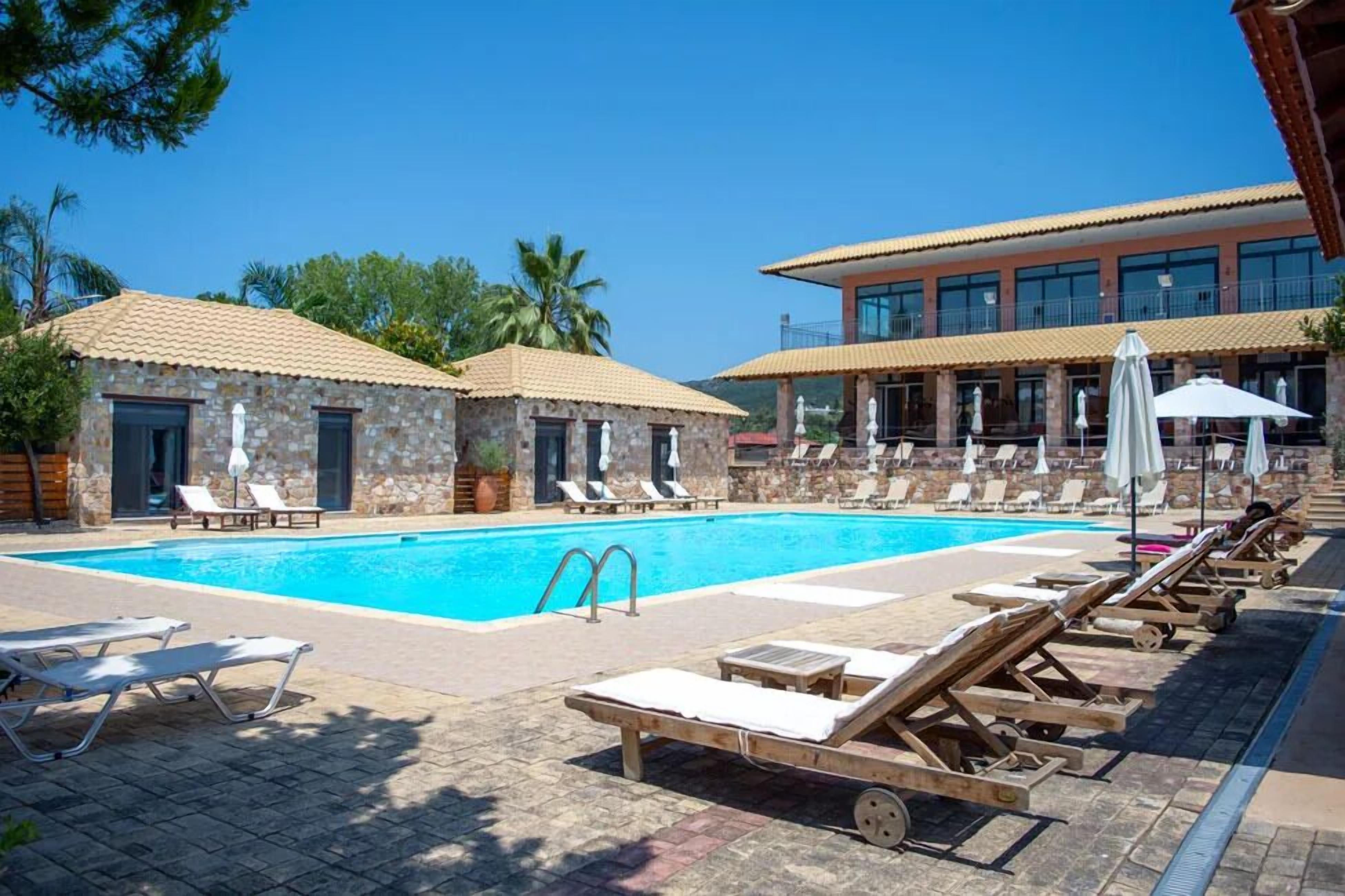 A swimming pool with lounge chairs and umbrellas, stone buildings with tiled roofs, and a clear blue sky.