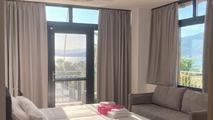 A hotel room with a bed, a sofa, and large windows offering a view of a balcony and mountains. - Almyra Holiday Village (Istiaia-Aidipsos)