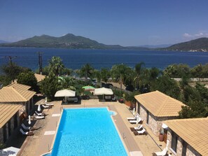 A pool area with lounge chairs and umbrellas, overlooking a body of water and hills. - Almyra Holiday Village (Istiaia-Aidipsos)