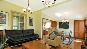 Sala de estar com um sofá de couro preto, uma poltrona bege e uma mesa de centro de madeira. Há duas luminárias pendentes, um quadro na parede e vista para a cozinha através de uma porta de vidro.