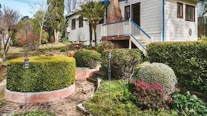 Uma casa de dois andares com varanda da frente, chaminé e um jardim bem cuidado, com arbustos variados e um cerca circular.