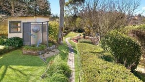 Um pequeno edifício térreo com porta e janela, rodeado por um jardim bem cuidado com diversas plantas e um caminho pavimentado.