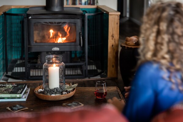 Une pièce chaleureuse avec un poêle à bois, une bougie dans un bocal en verre et un verre de vin rouge posé sur une table en bois.