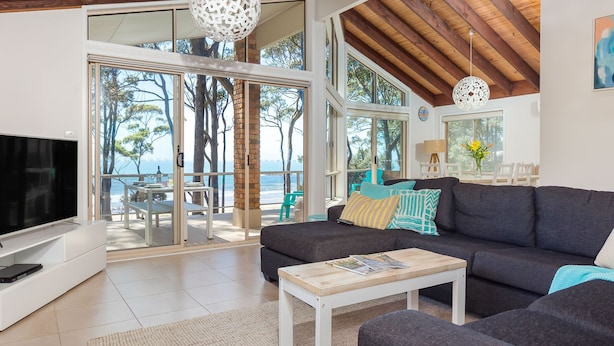 A modern living room with a large TV, a sofa, and a coffee table. There is a balcony with a view of trees and the sky. - Absolute Beachfront Penthouse | Panoramic Ocean Views | Batemans Bay Luxury (denhams beach)