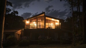 A modern two-story house with large windows and a well-lit interior, surrounded by trees and a garden. - Absolute Beachfront Penthouse | Panoramic Ocean Views | Batemans Bay Luxury (denhams beach)