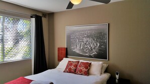 Ein Hotelzimmer mit Bett, Fenster mit Jalousien, einem Wandbild und einem Nachttisch mit Lampe.