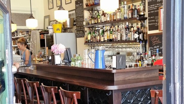 Eine Bar mit einer Holztheke, Barhocker und Regalen voller Flaschen. Eine Kreidetafel mit der Speisekarte ist an der Wand zu sehen.