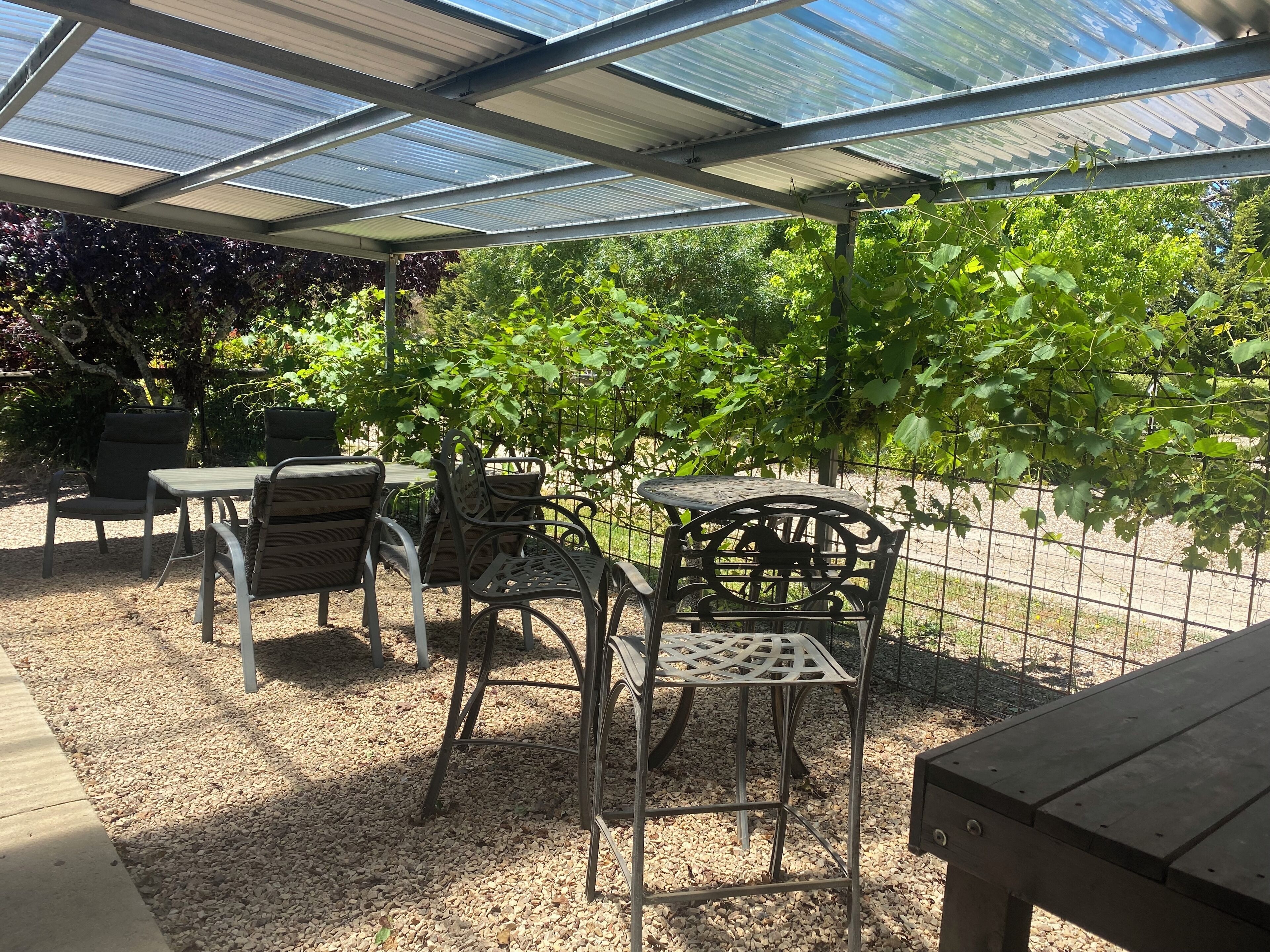 A covered outdoor seating area with metal chairs and a table, surrounded by a metal fence and greenery.