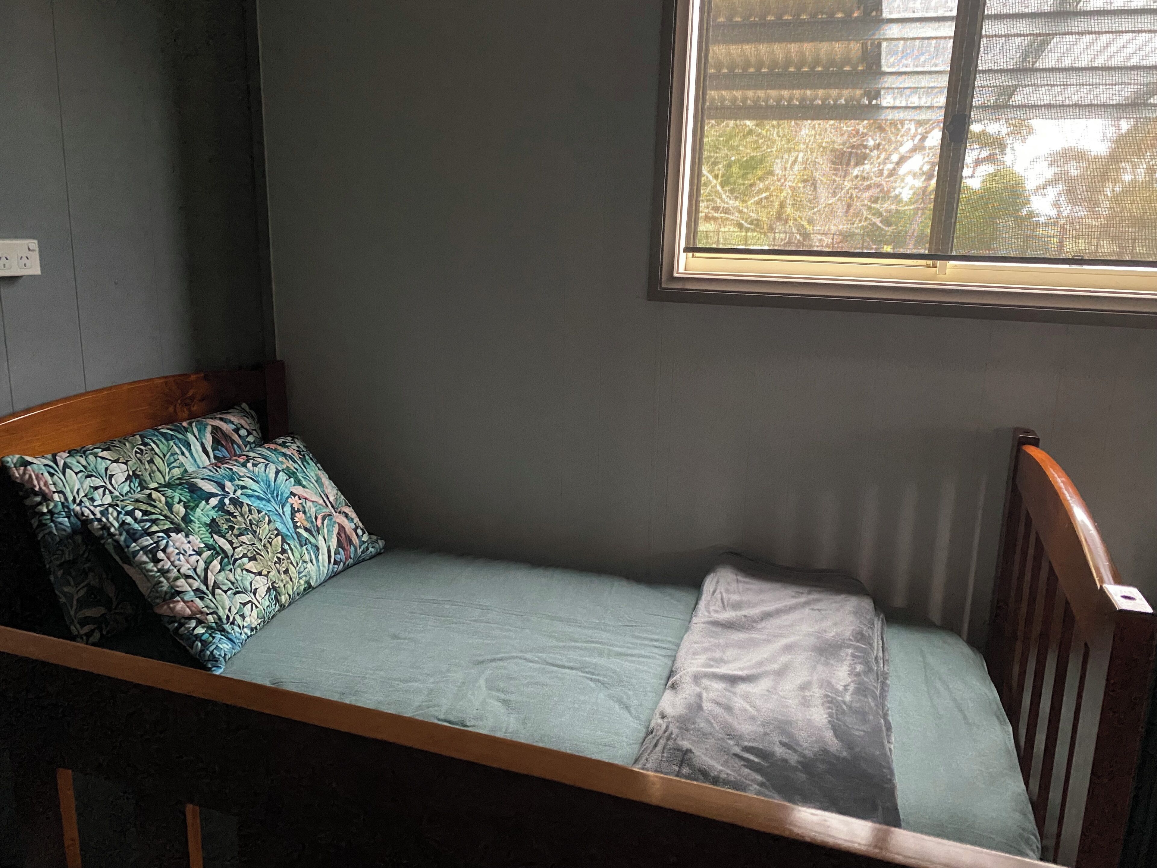 A single bed with a wooden headboard and a patterned pillow, a window with a view of trees, and a wall socket.