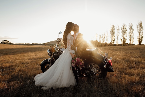 Un couple s’embrasse sur une moto, dans un champ, avec un coucher de soleil en arrière-plan.