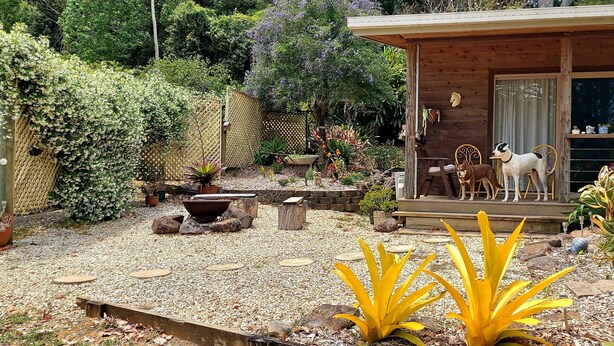 Una cabaña de madera con un porche, rodeada por un jardín con diversas plantas y dos perros.