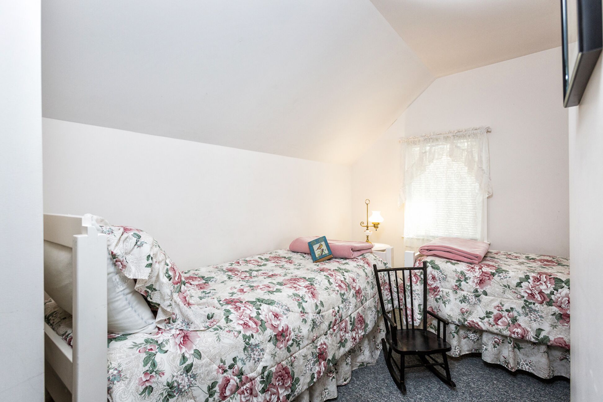 A bedroom with two single beds, a nightstand with a lamp, a chair, and a window with a sheer curtain.