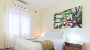 A hotel room with a bed, a nightstand, a painting of fruit on the wall, and a window with curtains.