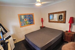 Una camera da letto con un letto, una ventola a soffitto, uno specchio fissato al muro e un comodino con una lampada.