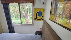 Une chambre à coucher avec un lit, une fenêtre donnant sur des arbres, un tableau accroché au mur et une petite table sur laquelle se trouve un objet rouge.