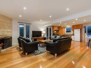 A spacious living area with a fireplace, leather sofas, a flat-screen TV, and a kitchen with wooden cabinets.