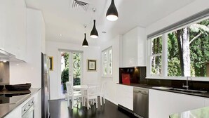 A modern kitchen with white cabinetry, a black countertop, and a stainless steel dishwasher. There is a dining area with white chairs and a table, and a window with a view of trees.