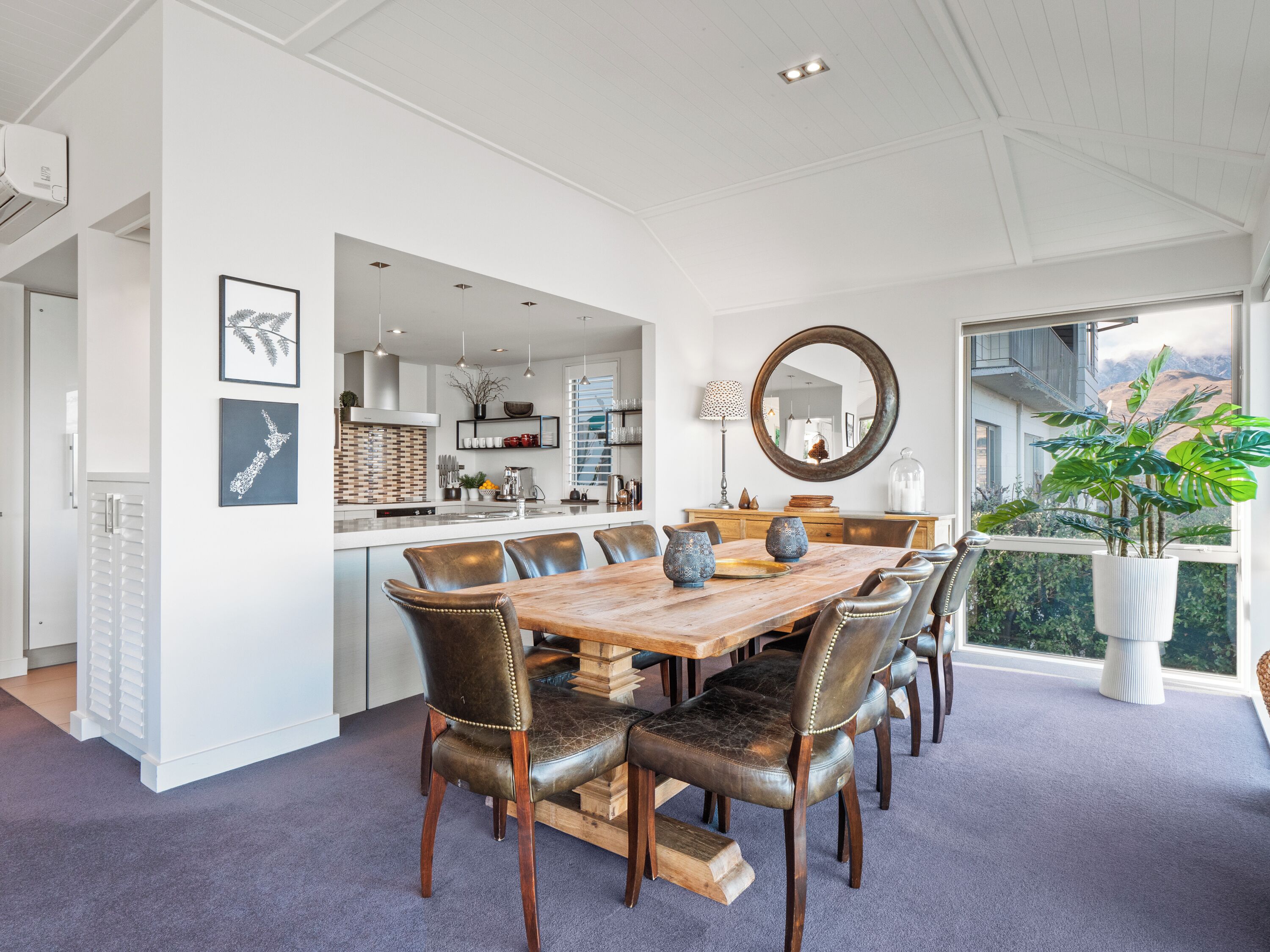 A spacious dining area with a wooden table and leather chairs, a kitchen in the background, and a large window with a view of greenery.