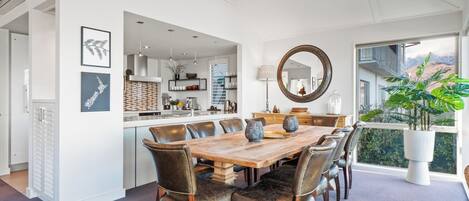 A spacious dining area with a wooden table and leather chairs, a kitchen in the background, and a large window with a view of greenery.