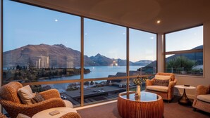 A spacious room with large windows offering a view of mountains and a lake. There are wicker chairs, a round coffee table, and a small side table. - Panorama, Queenstown (Queenstown Hill)
