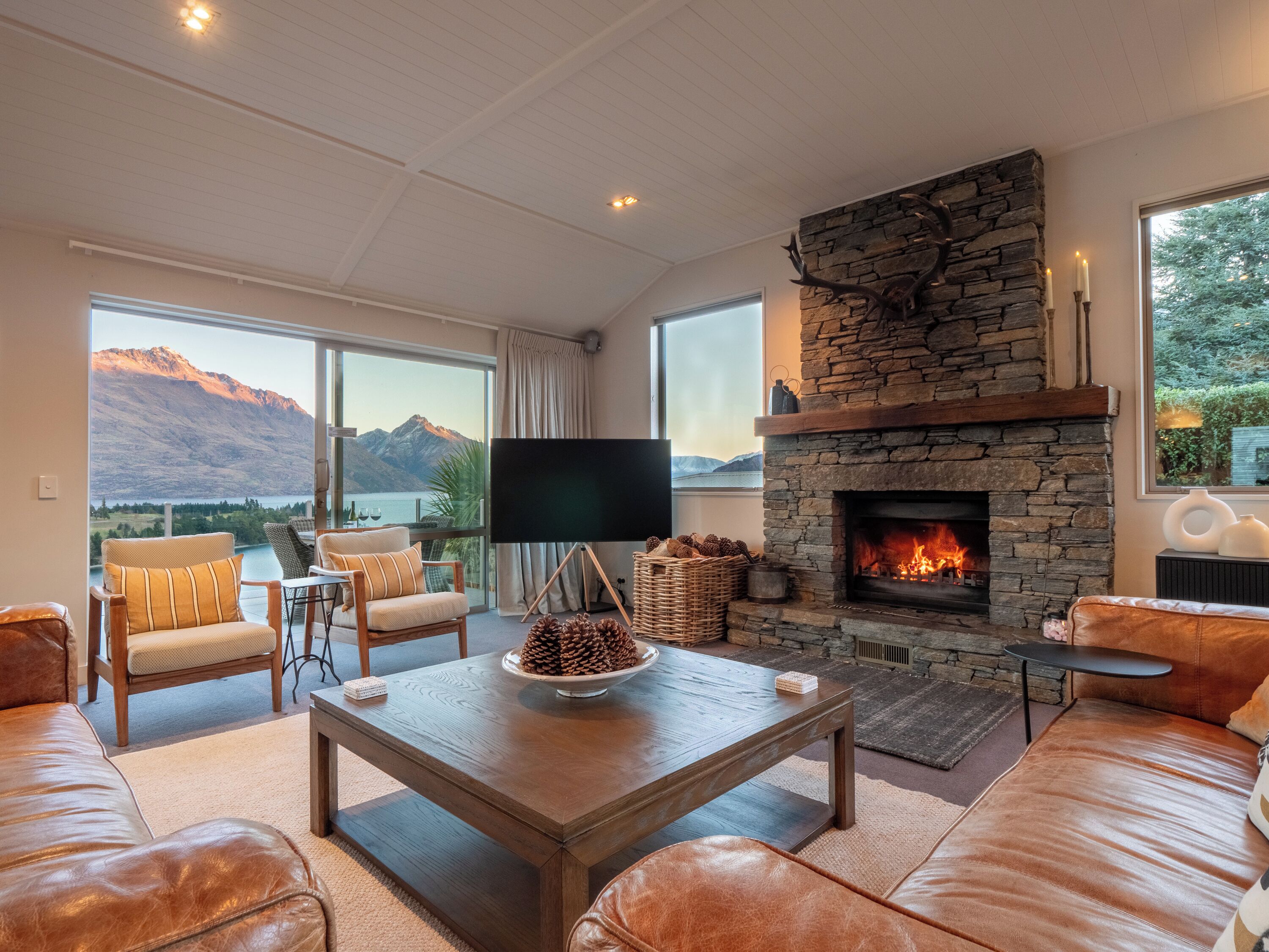 A spacious living room with a stone fireplace, leather sofas, a large flat-screen TV, and a view of mountains and a lake.