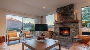 A spacious living room with a stone fireplace, leather sofas, a large flat-screen TV, and a view of mountains and a lake. - Panorama, Queenstown (Queenstown Hill)