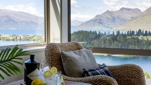 A wicker sofa with cushions and a tray with a bottle, glasses, and lemons by a window overlooking a lake and mountains. - Panorama, Queenstown (Queenstown Hill)