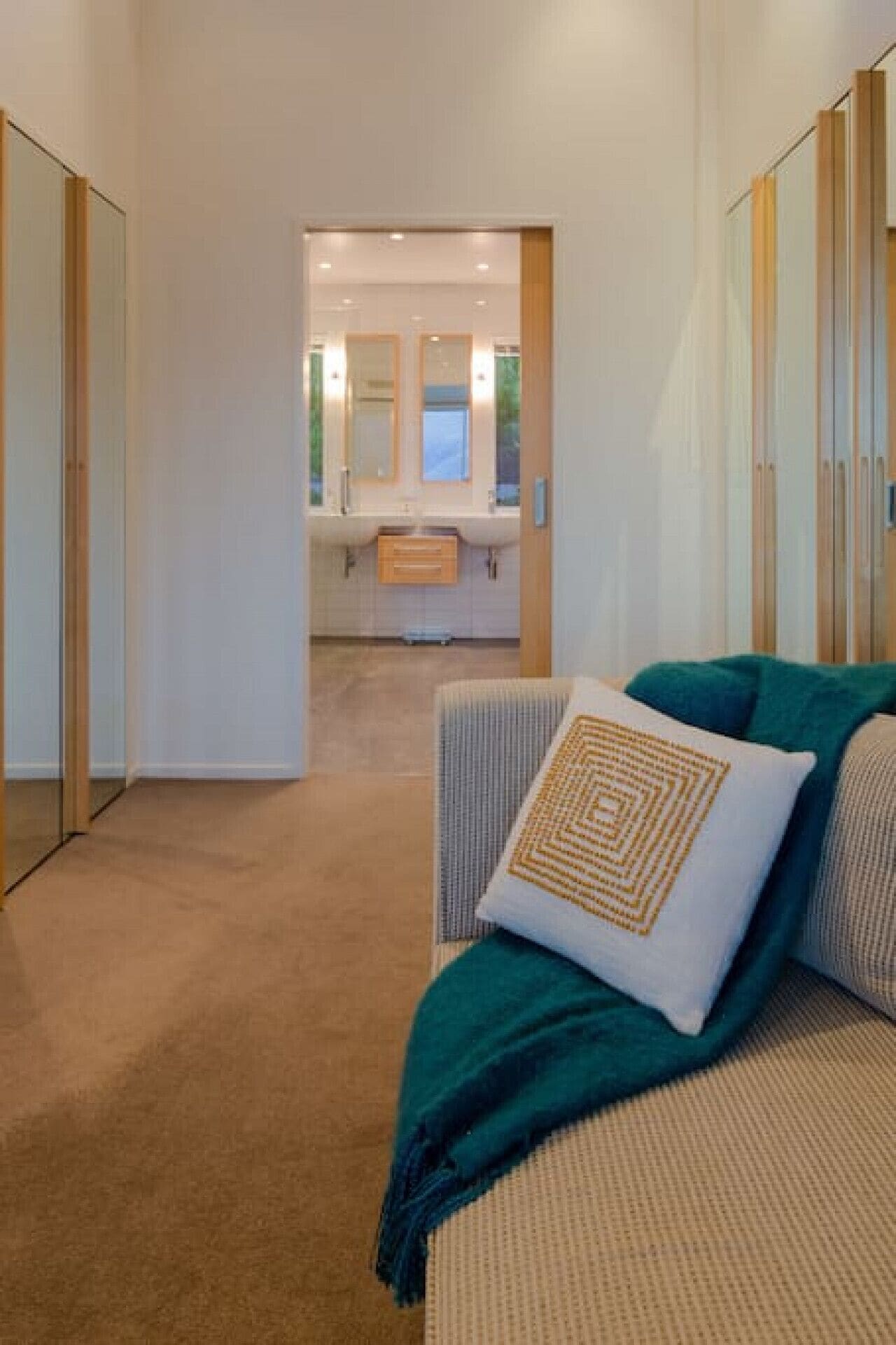 A hallway leading to a bathroom with a sink and mirrors. There is a beige sofa with a patterned pillow and a green throw blanket.