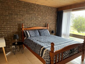 A bedroom with a brick wall, wooden bed frame, patterned bedding, a bedside table with a lamp, and a window with curtains. - Family Beach Home Dromana -Relax and take in the views of the bay (Dromana)