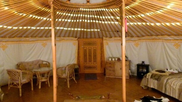 Habitación con un dosel de madera central, una cama con edredón a rayas, una pequeña mesa con sillas y una alfombra decorativa.