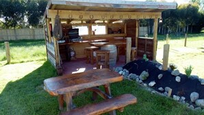 Una cocina rústica al aire libre con una mesa y bancos de madera, rodeada por un muro de piedra y follaje.