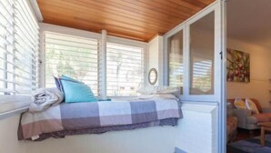 A bedroom with a bed against a wall, a window with blinds, a door leading to another room, and a small rug on the floor.