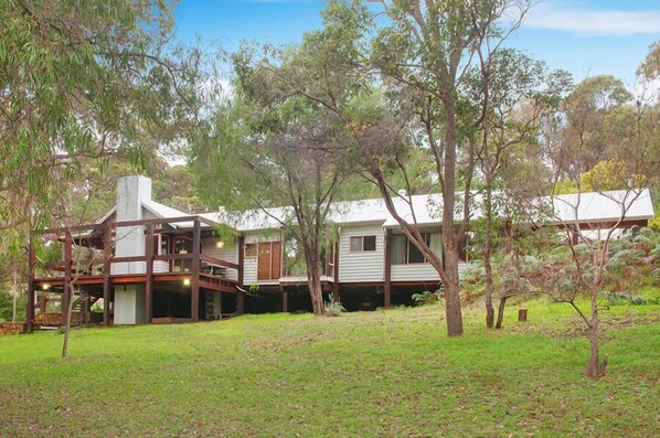 A two-story house with a deck and a chimney, surrounded by trees and a grassy yard. - Wilyabrup River Retreat - A gorgeous house on a private vineyard, wood fire (Wilyabrup)