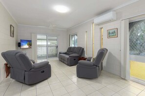 A spacious living room with grey reclining chairs, a flat-screen TV, and a sliding glass door leading to a balcony.