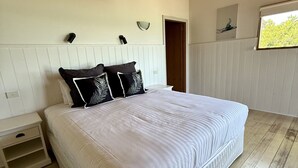 A bedroom with a large bed, white walls, a wooden floor, a nightstand, and a window with a view of trees.
