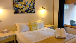 Cama bem arrumada com lençóis brancos e uma toalha dobrada, mesa de cabeceira com caixa de lenços, pintura na parede e janela com vista para o exterior.