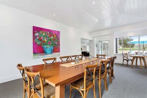 A dining area with a wooden table and chairs, a vibrant painting on the wall, and a view through large windows.
