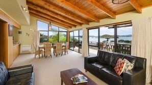 Un vaste espace de vie doté d’un plafond en bois, d’un canapé en cuir noir, d’une table à manger avec des chaises et de grandes fenêtres offrant une vue sur la mer et les collines.