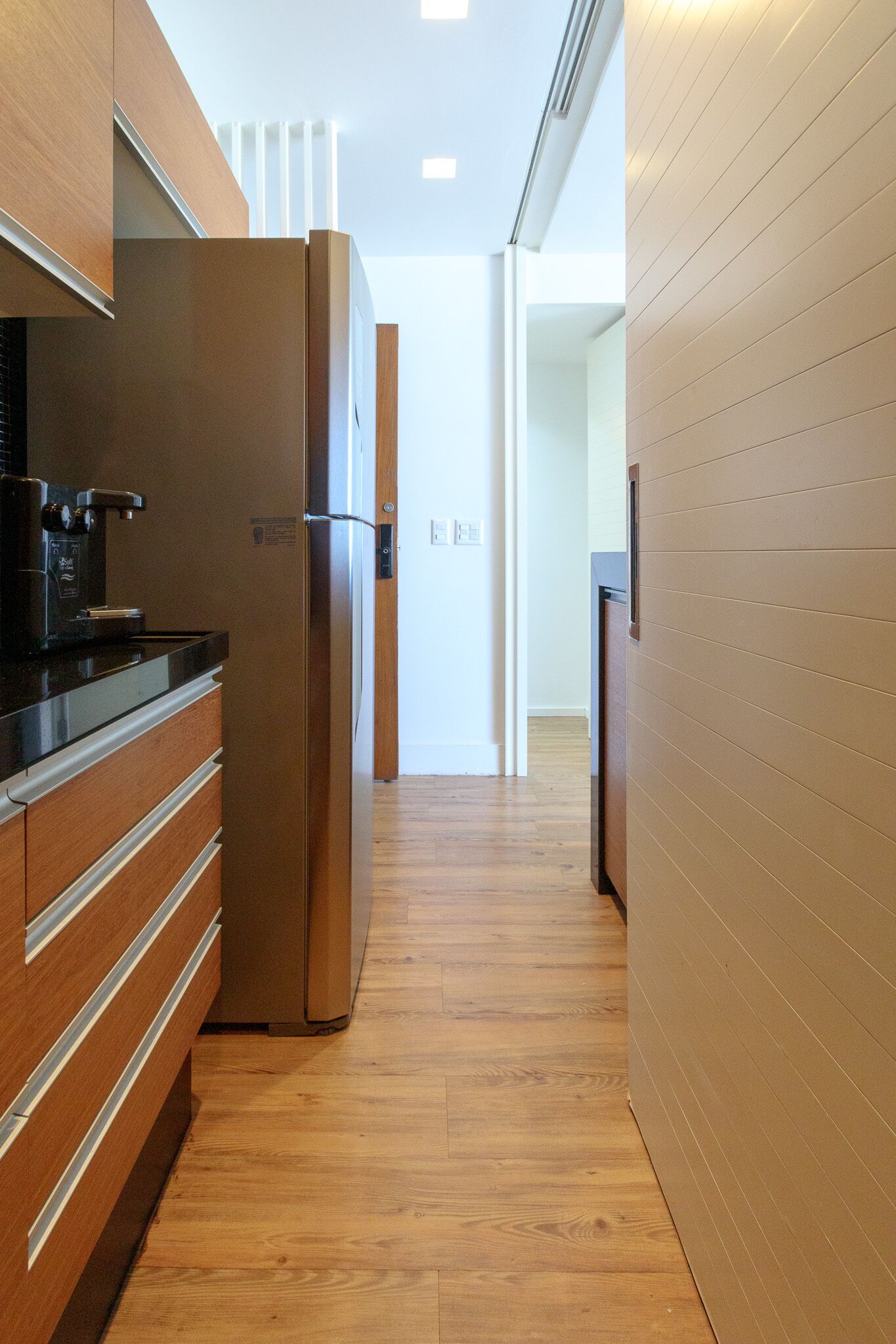 A narrow hallway with wooden flooring, a stainless steel refrigerator, and a coffee maker on the left.