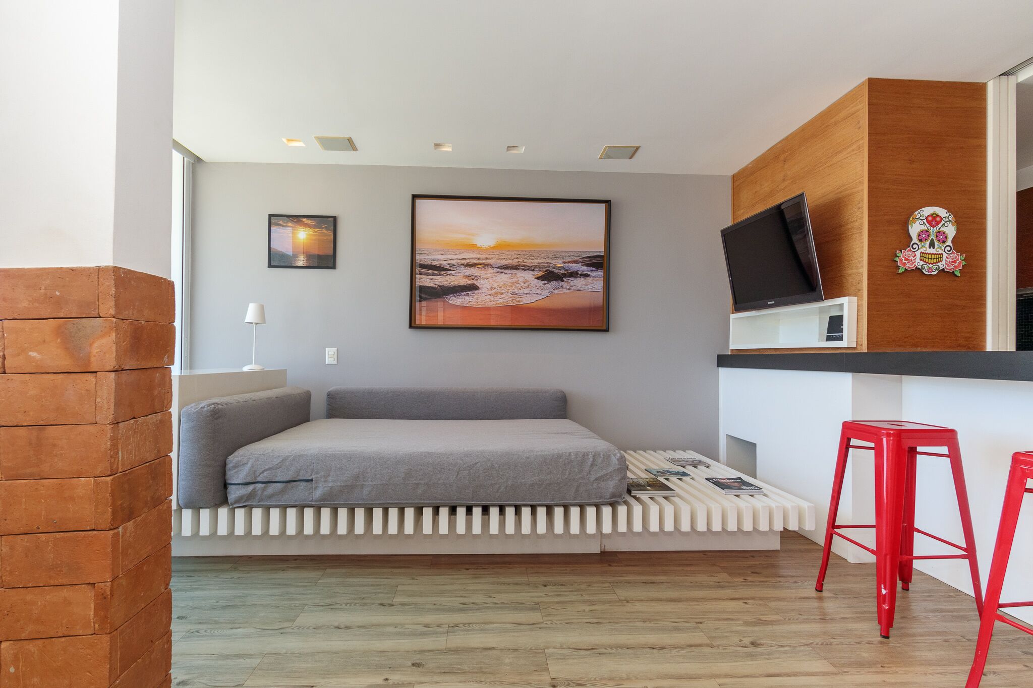 A modern room with a bed, a TV, a red stool, and a picture of a sunset.