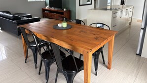 Un salon moderne avec un canapé en cuir foncé, une table à manger en bois avec des chaises, un téléviseur posé sur un meuble en bois, et un coin cuisine doté de meubles blancs et d’un réfrigérateur en acier inoxydable.