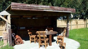 An outdoor wooden structure with a round table and chairs, surrounded by a grassy area.