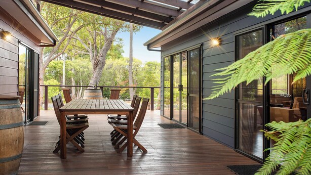 Une terrasse extérieure avec une table et des chaises en bois, une pergola au-dessus et une vue sur des arbres et un bâtiment en arrière-plan.