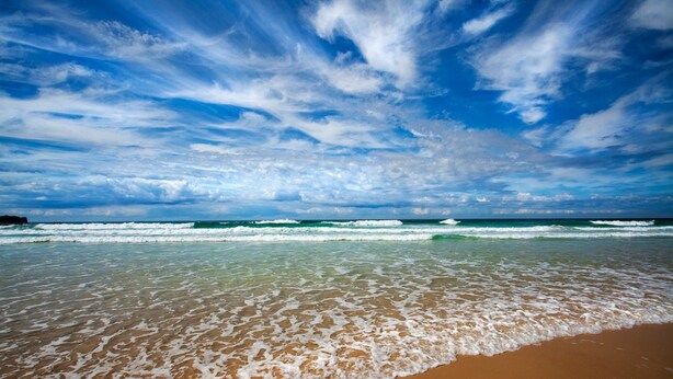 A beach with waves, clear skies, and a distant rocky outcrop.