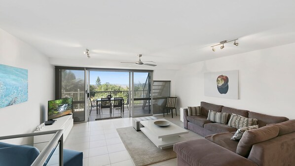 A modern living room with a sofa, coffee table, and a TV. There is a balcony with a view of greenery and a staircase. - Ocean Terraces - Split Level Apartment At The Beach - Ocean Views, Rooftop Terrace - 3 bed, 2 bath (SUNSHINE BEACH)