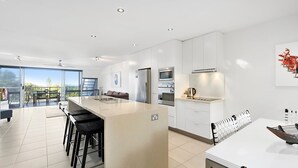 A modern kitchen with a central island, a dining area with a white table and black chairs, and a large sliding glass door leading to an outdoor balcony.