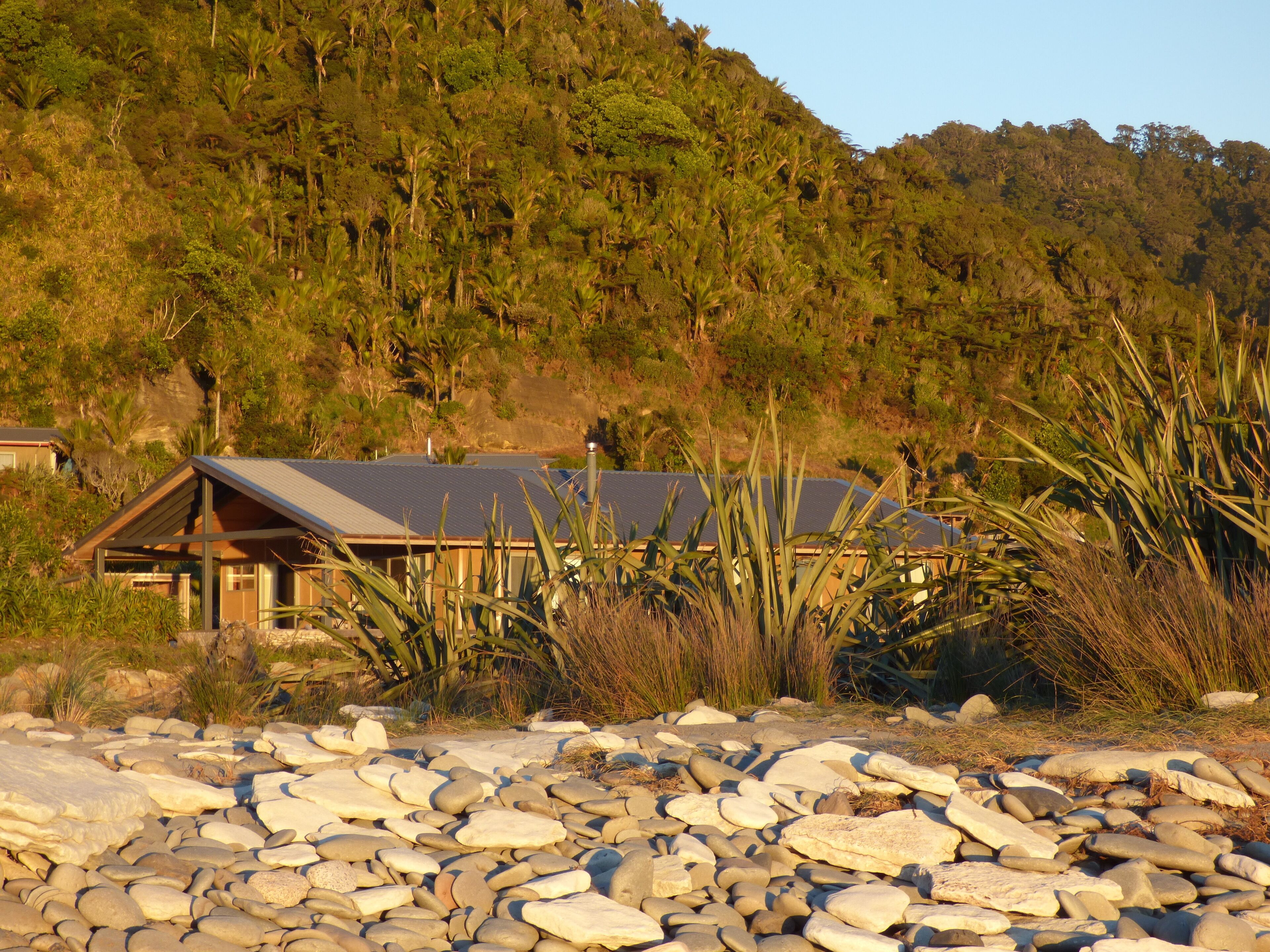 A house with a sloped roof, surrounded by a rocky beach and dense vegetation.