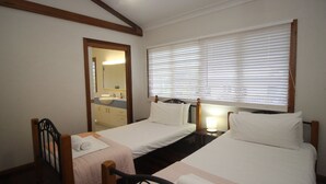 A bedroom with two single beds, a wooden headboard, a nightstand with a lamp, and a bathroom visible through an open door. - Magnetic Dreams Unit 1 (ARCADIA)
