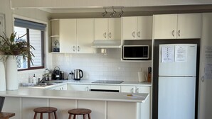 Une cuisine moderne avec des meubles blancs, un réfrigérateur en acier inoxydable, un four à micro-ondes et un évier. On y trouve trois tabourets en bois et une plante en pot près de la fenêtre.